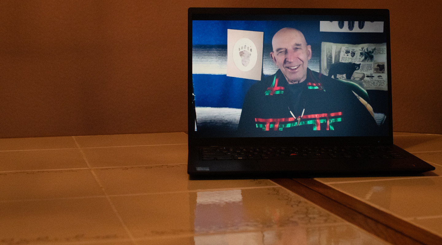 photo of Joseph Bruchac, an indigenous man wearing a black shirt with red and green ribbons, speaking on a computer screen