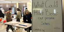 Three people look at exhibits laid out on tables near a sign that reads "What could have been: our possible selves."