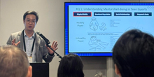 Johnny Cho presents to a group of listeners in front of a video screen.