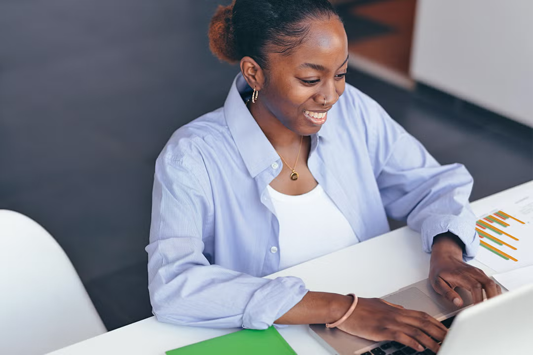 Smiling woman in a light blue shirt, focused and engaged in her work, types on a laptop at a desk with charts and a green folder.