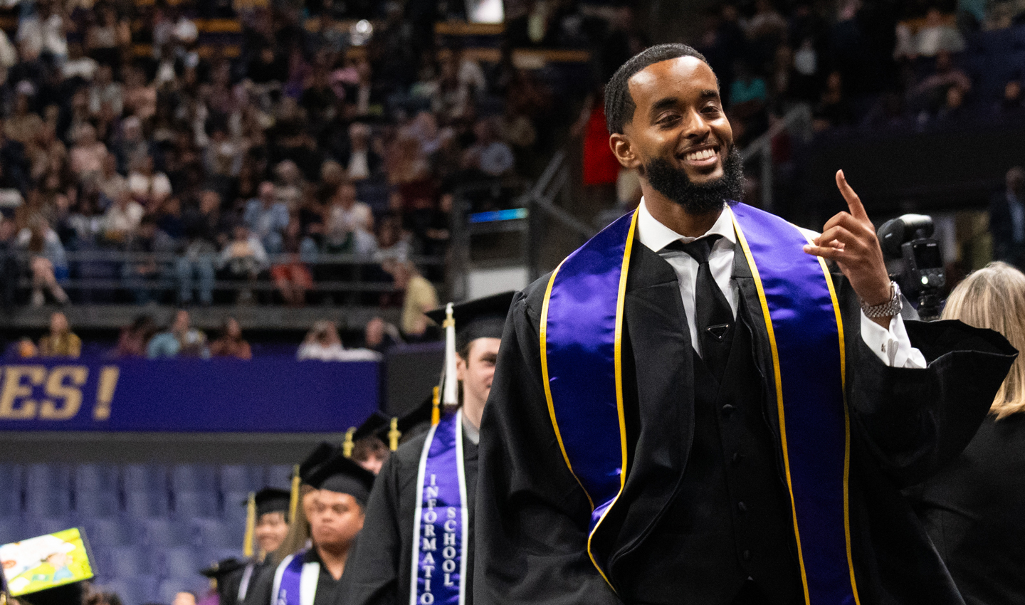 A man in graduation clothing smiles and gestures with his hand.