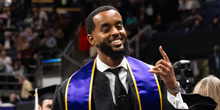 A man in graduation clothing smiles and gestures with his hand.