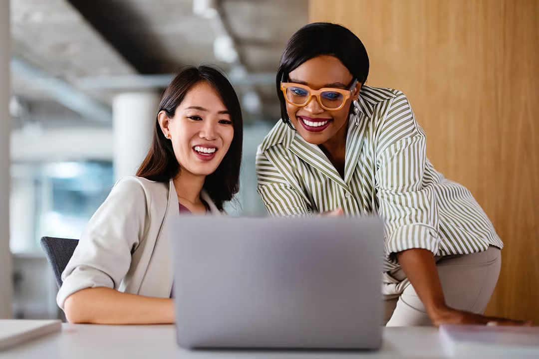 Two professional women smile while looking at a laptop screen in a modern office setting.