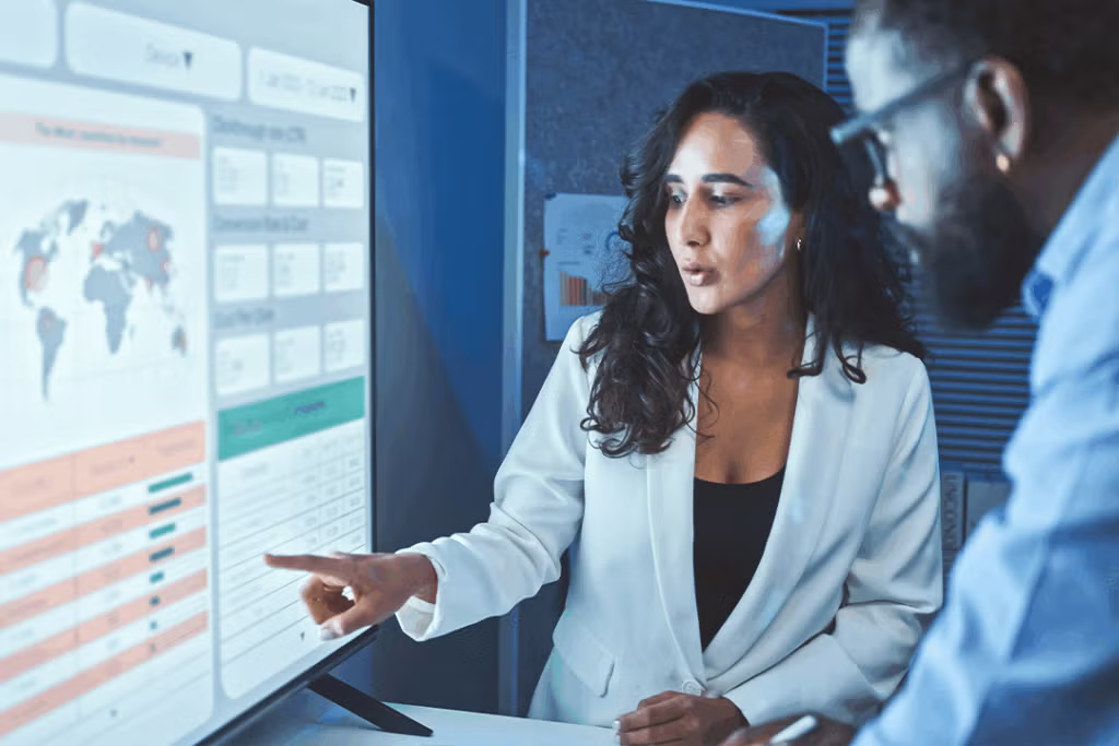 A woman in a white blazer points at a large screen displaying data graphs and a world map, while a man observes attentively beside her.