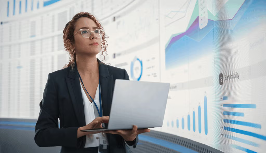 A young female professional holds an open laptop computer while considering a massive screen displaying data graphics.