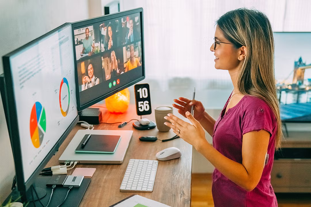 A young woman participates in a video conference call at a desk with multiple monitors displaying charts and meeting participants.