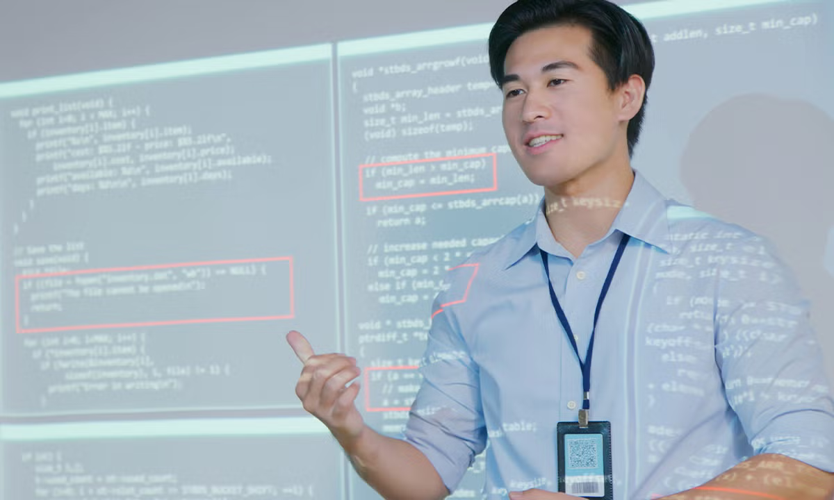 A presenter stands in front of a large screen displaying lines of computer code, with some sections highlighted in red, gesturing as he explains a programming concept.