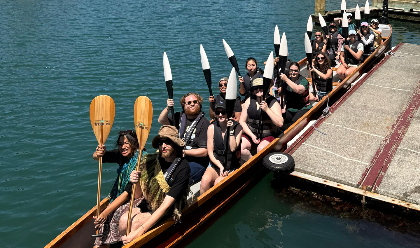 Students hold paddles up while seated in a canoe.