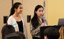 Two students present in front of a room of visitors.