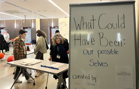 Three people look at exhibits laid out on tables near a sign that reads "What could have been: our possible selves."