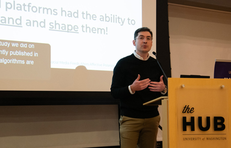 A man speaks at a lectern in front of a screen showing a projection.
