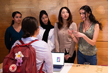 A row of women behind a table with another person in the foreground. One of the women behind the table is explaning the team's project.