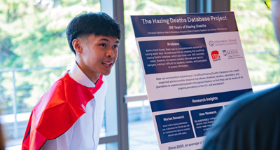 A young man wearing a cape speaks in front of a poster display.
