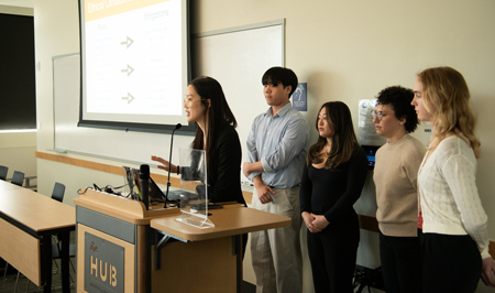 A group of 5 students presents at the front of a room.