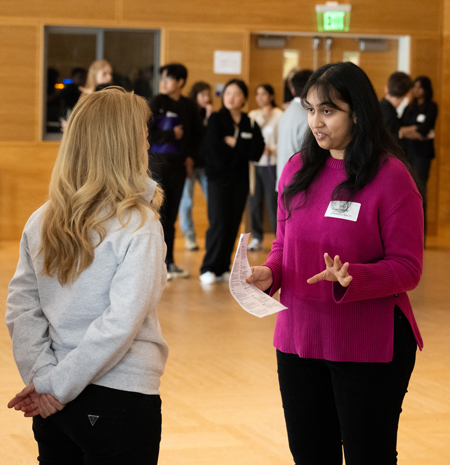 Two women speak while standing. One is holding a piece of paper.