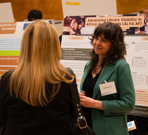 Renee Lynch speaks with a person with their back to the camera while standing in front of reserach posters.