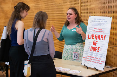 A woman gestures while speaking next to a poster reading "Library of Hope."